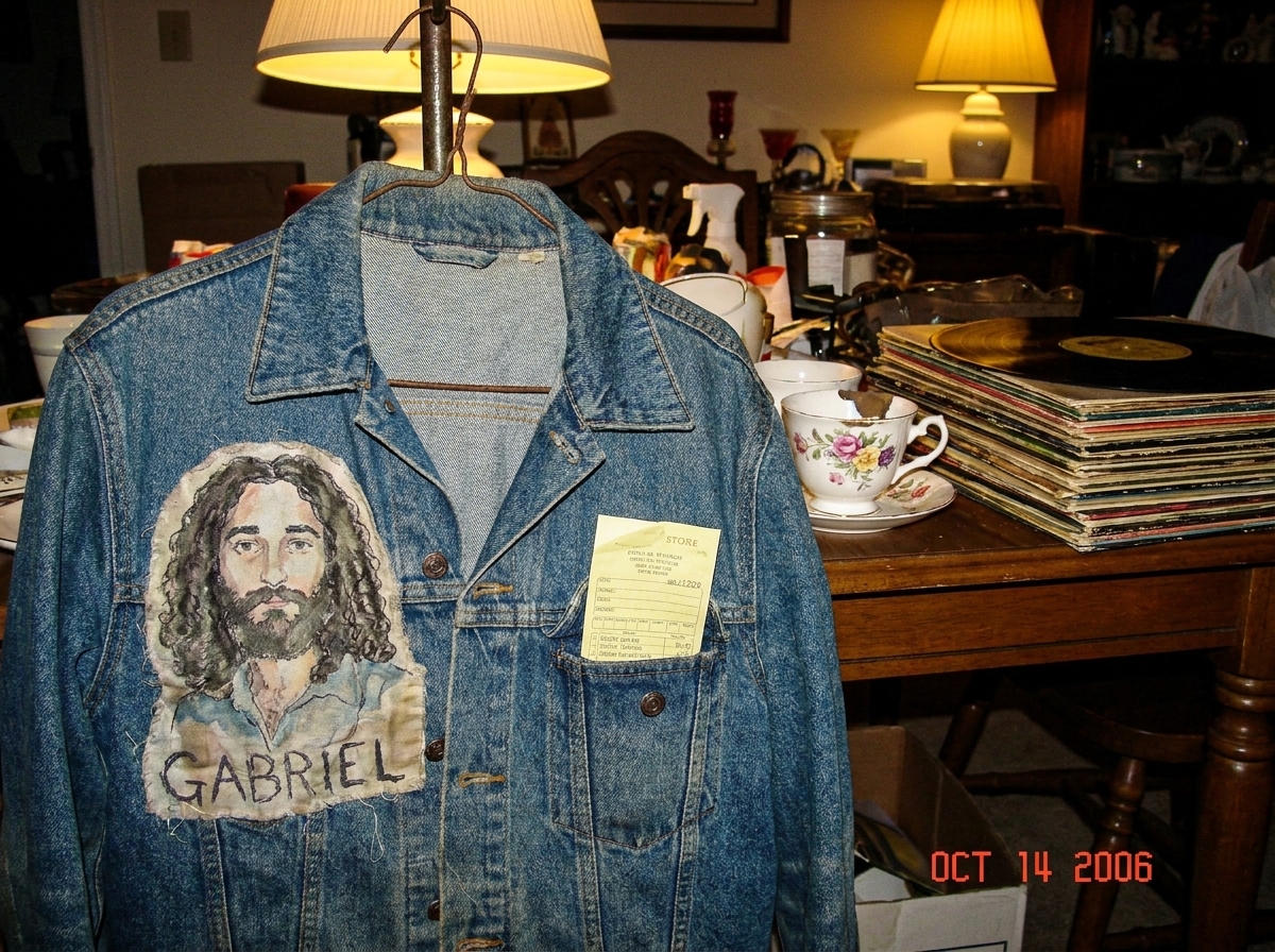 A faded denim jacket hanging on a wire hanger at a yard sale, a yellowed store receipt peeking from one of the pockets. The jacket is worn at the elbows and cuffs, with a small embroidered patch on the left chest that reads 'GABRIEL' in uneven stitching. The background is a cluttered yard sale table