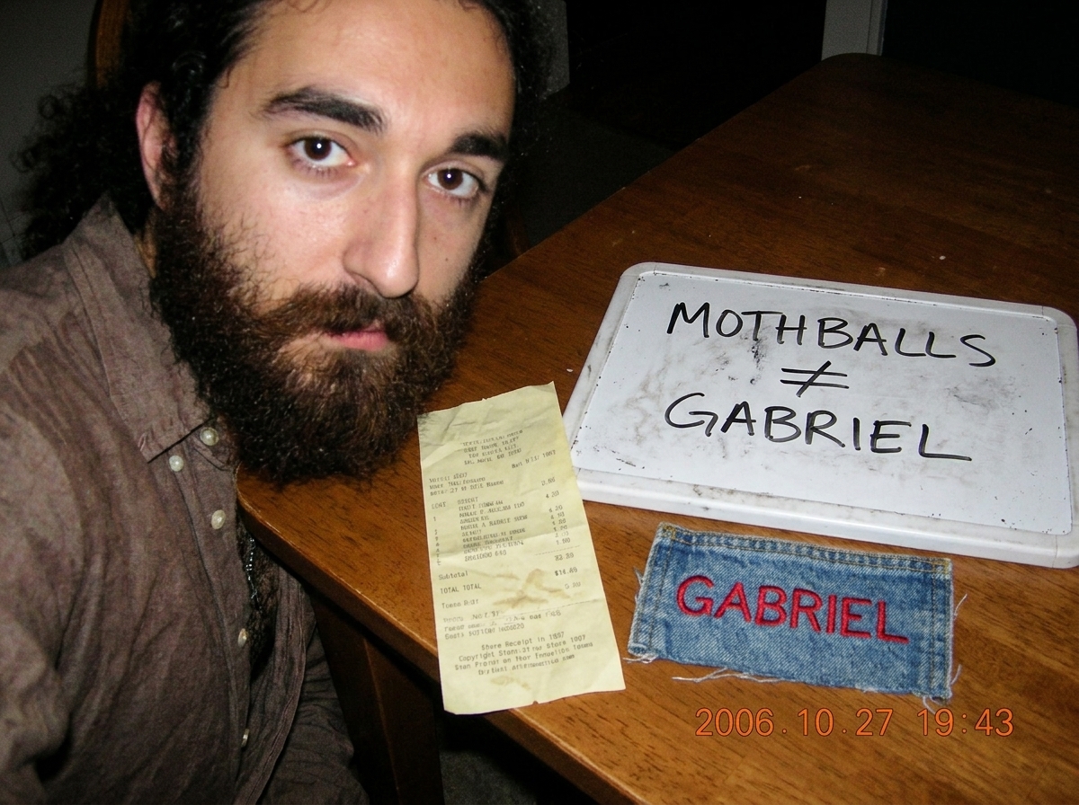 A close-up photo of a crumpled yellowed store receipt from 1997 placed next to a whiteboard where someone has written 'MOTHBALLS != GABRIEL' in black marker. A single faded denim jacket patch embroidered with 'GABRIEL' sits beside the receipt.