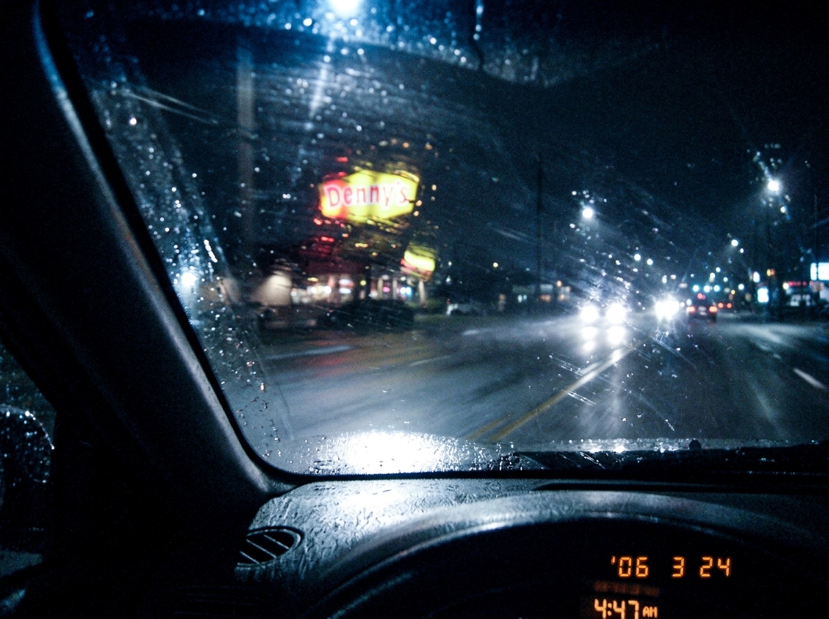 A blurry, dark photo taken from inside a car at night, showing a Dennys sign in the distance with the letters partially obscured by rain on the windshield. The timestamp in the corner reads 4:47 AM but it's too pixelated to verify.