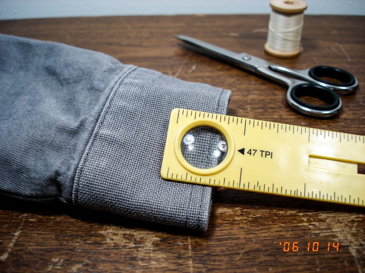 A close-up photo of a gray fabric cuff from 2003, laid flat on a wooden sewing table next to a yellow plastic sewing gauge with a magnifying lens. The gauge is positioned over the fabric weave, showing the measurement marking at 47 threads per inch. The fabric has a visible grid pattern. In the back