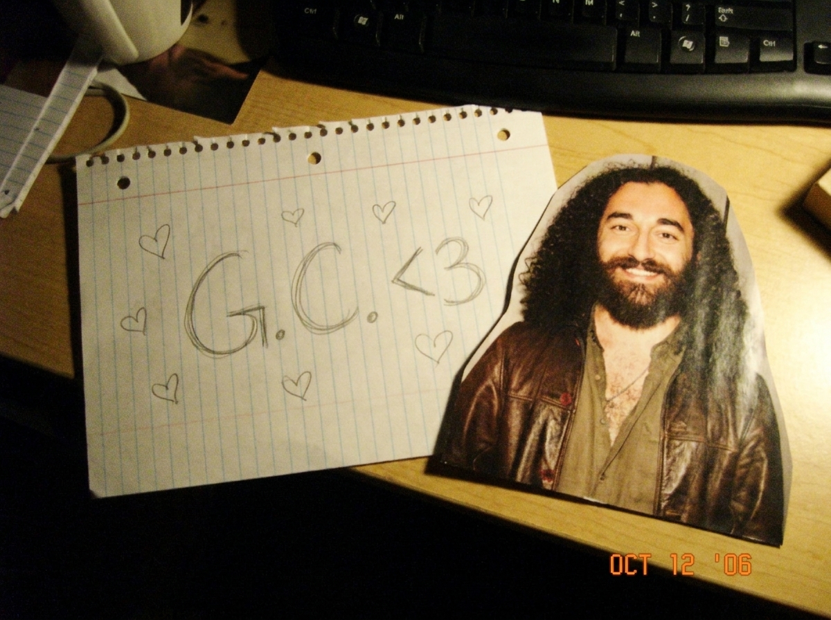 A handwritten note on notebook paper that says 'G.C. <3' doodled in pencil with little hearts around it, sitting on a bedroom desk next to a magazine photo of Gabriel