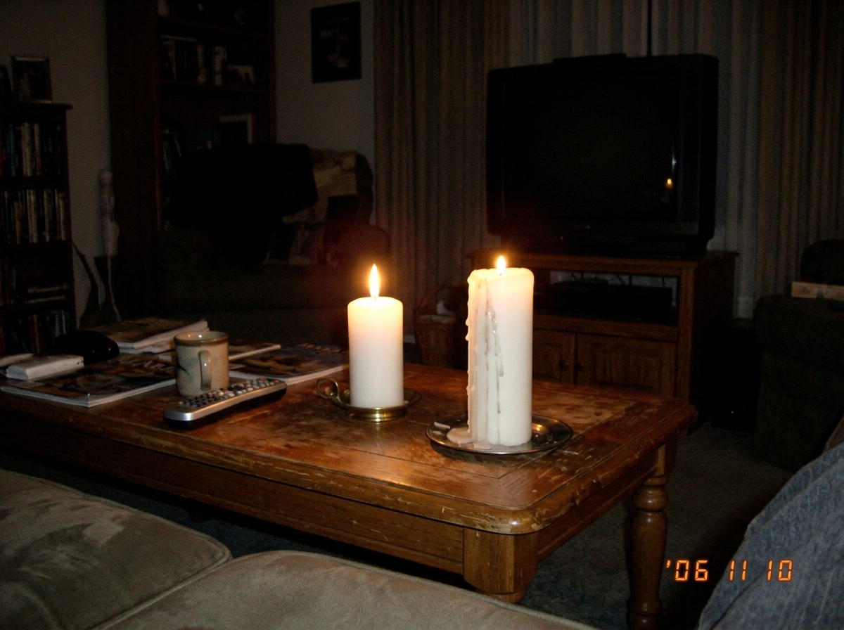 A blurry, dimly lit photo taken from a couch showing two white pillar candles burning on a coffee table. The TV in the background is dark and off. One candle has a slight drip down the side. The photo has a grainy, low-light digital camera quality from 2006.
