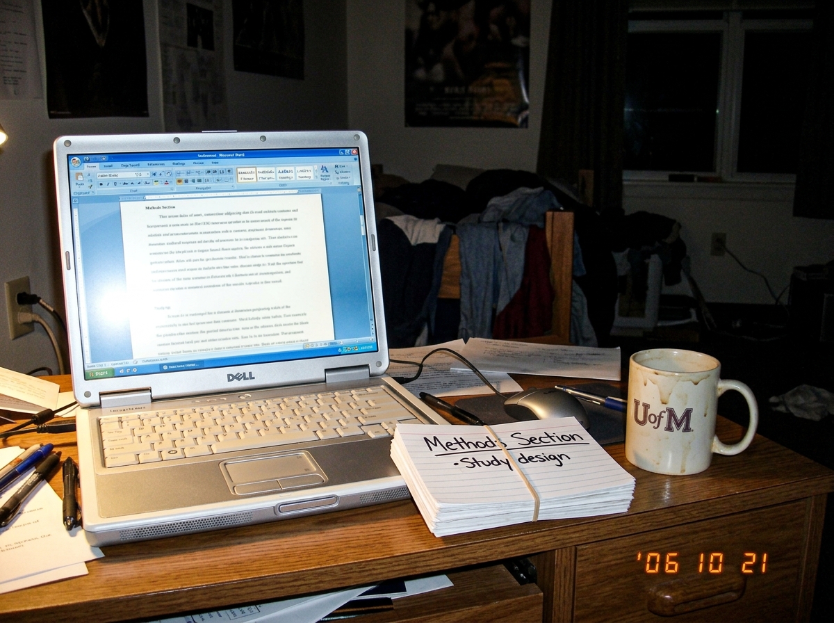 A dimly lit bedroom desk at night showing a single laptop screen glowing next to a stack of index cards labeled 'Methods Section' and a half-empty mug of coffee