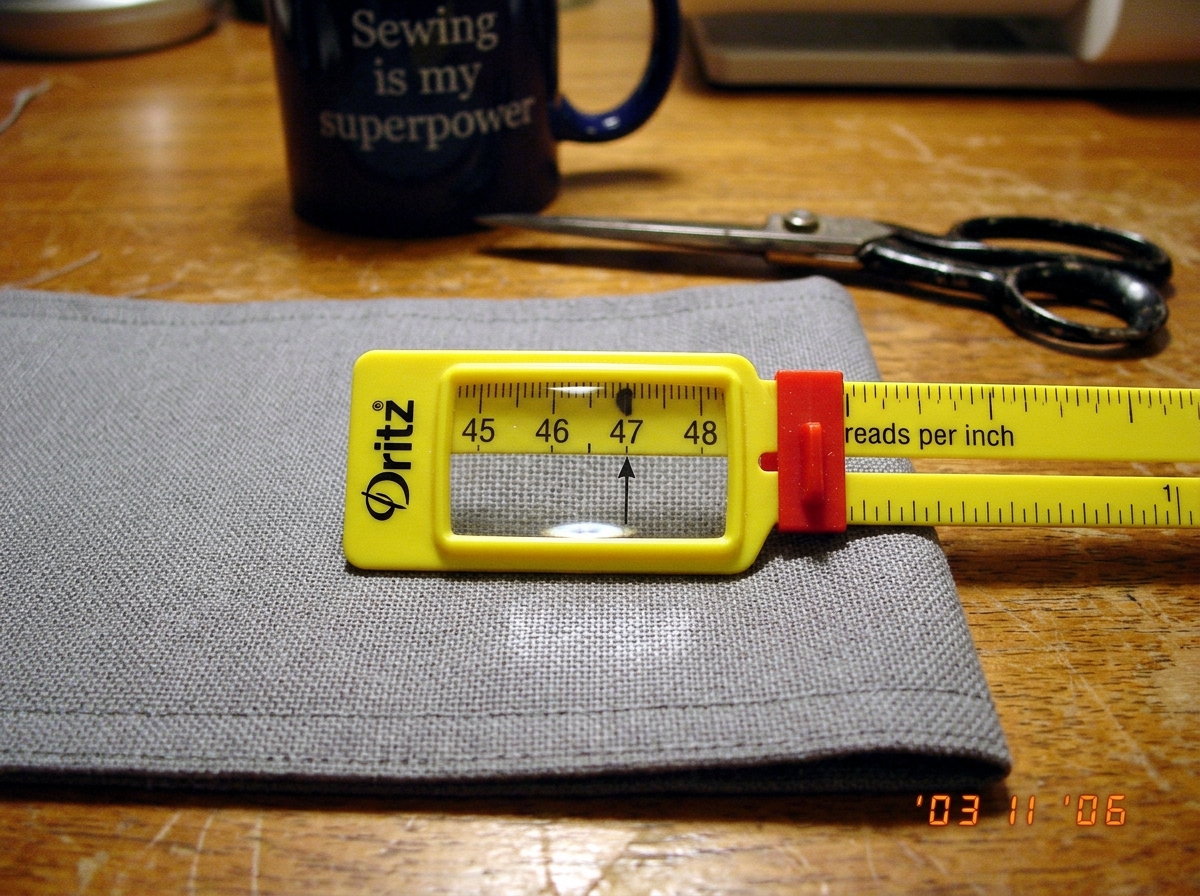 A close-up photo of a gray fabric cuff from 2003, laid flat on a wooden sewing table next to a yellow plastic sewing gauge with a magnifying lens. The gauge is positioned over the fabric weave, showing the measurement marking at 47 threads per inch. The fabric has a visible grid pattern. In the back