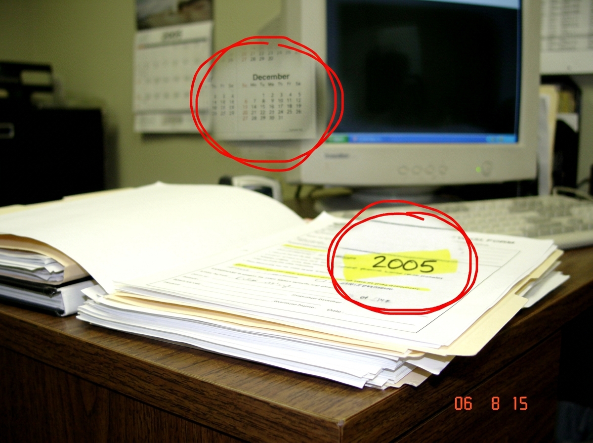 A blown-up crop of a desk photo ElderProphet07 posted in August 2006 showing a correction photo. In the background reflection on a monitor or glass surface, a 2005 wall calendar is faintly visible with December visible. On the foreground document, highlighter bleed-through is visible from the opposi