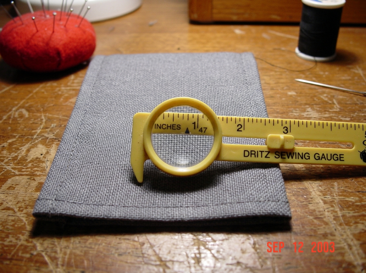 A close-up photo of a gray fabric cuff from 2003, laid flat on a wooden sewing table next to a yellow plastic sewing gauge with a magnifying lens. The gauge is positioned over the fabric weave, showing the measurement marking at 47 threads per inch. The fabric has a visible grid pattern. In the back