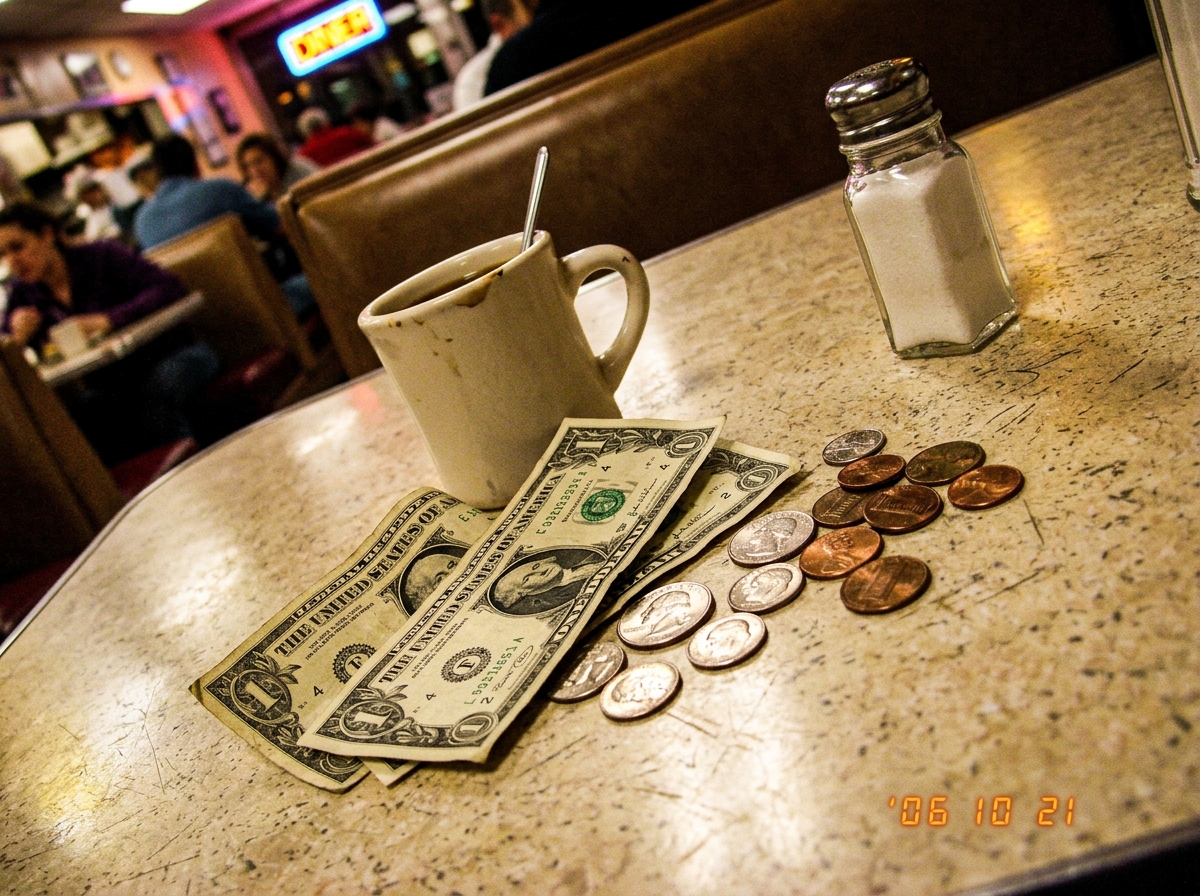 A close-up of a diner booth 4 table with a $4.71 tip in singles scattered across a Formica surface, next to a coffee cup and a salt shaker. The lighting is harsh, casting sharp shadows, and the angle is slightly tilted to emphasize the discrepancy in coin denominations. The background is blurred, fo