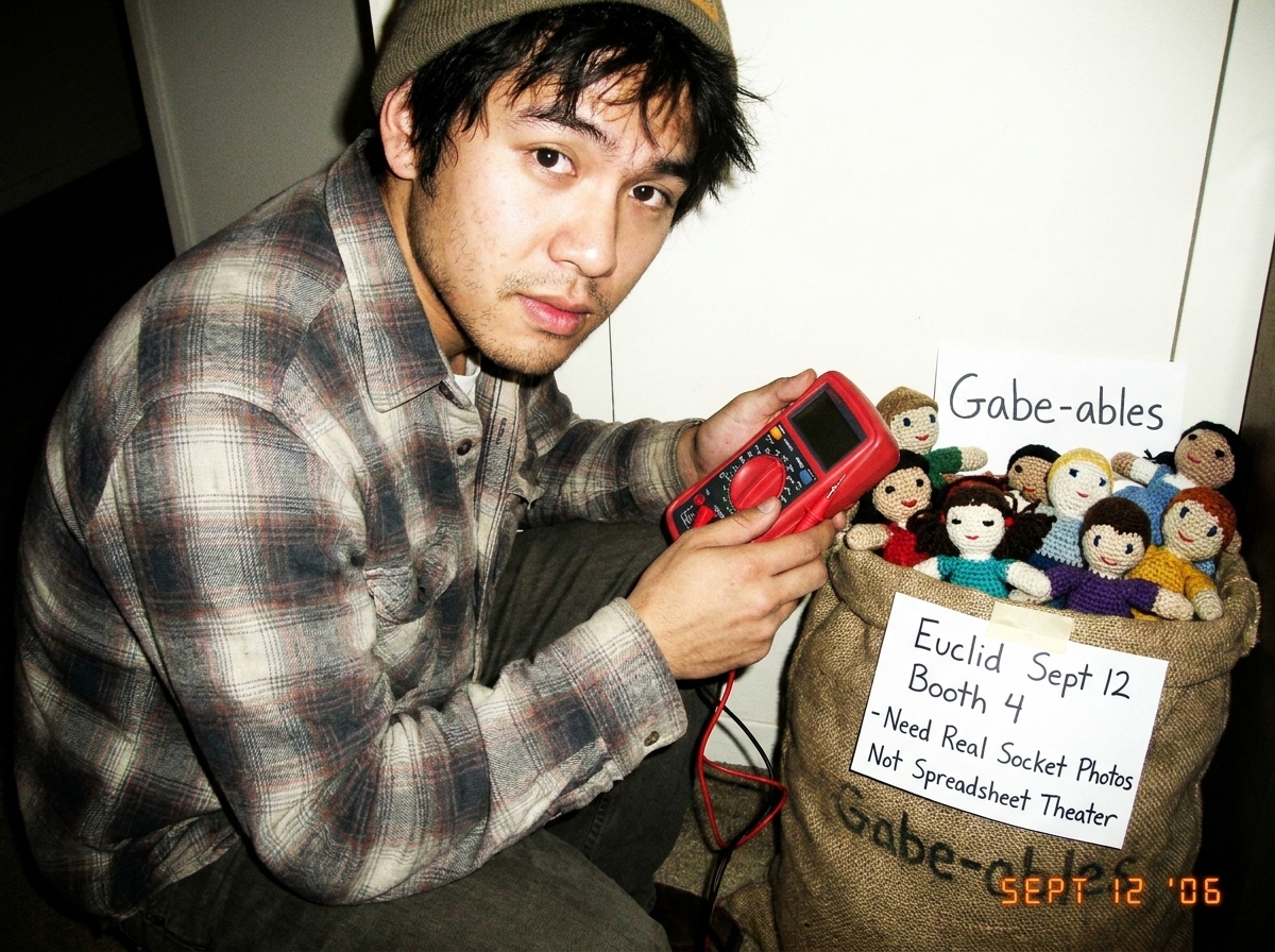 A photo of a digital multimeter probe resting next to a burlap sack of Gabe-ables crocheted dolls, with a handwritten note taped to the sack that reads 'Euclid Sept 12 Booth 4 - Need Real Socket Photos Not Spreadsheet Theater'