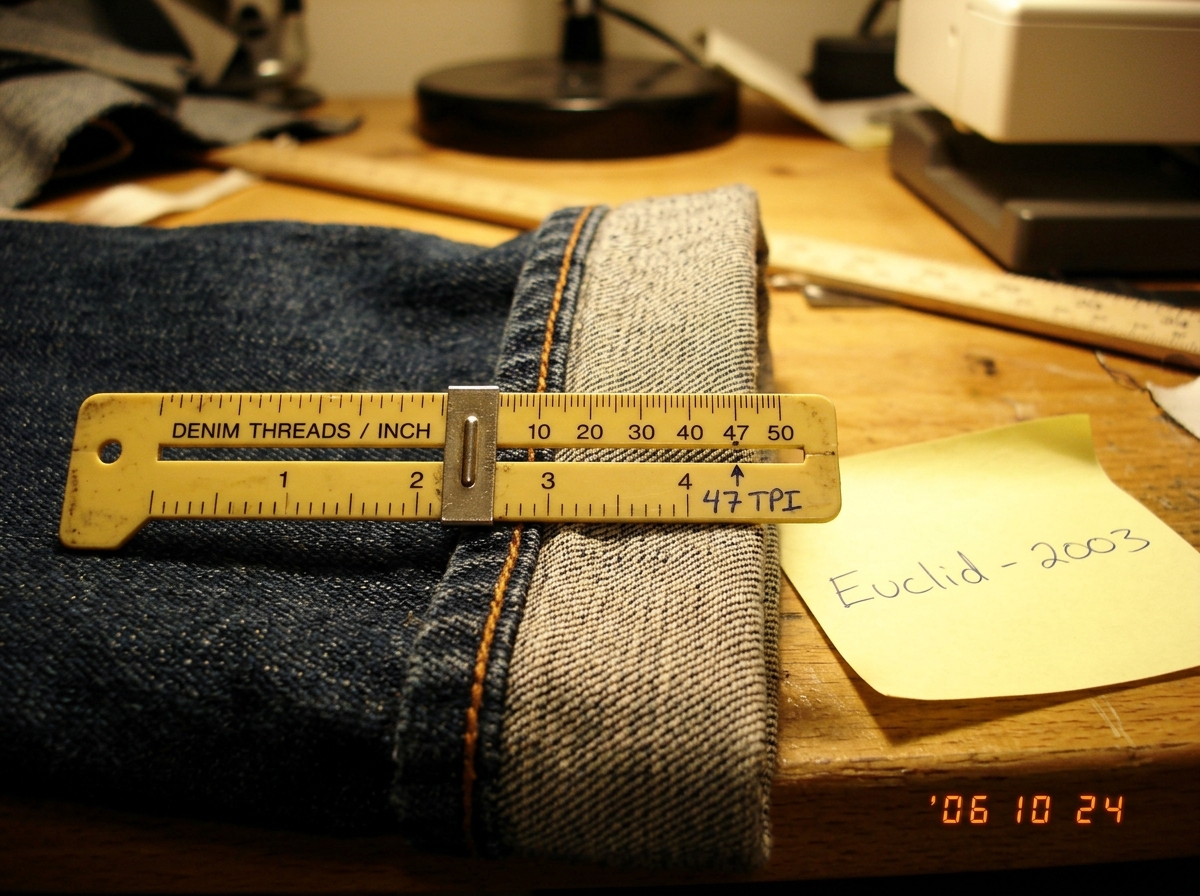 A close-up photograph of a denim cuff showing the woven edge, with a yellow sewing gauge laid across it measuring 47 threads per inch. The photo is slightly out of focus and the lighting is warm from a desk lamp. A handwritten sticky note at the edge reads 'Euclid - 2003' in blue ballpoint pen.