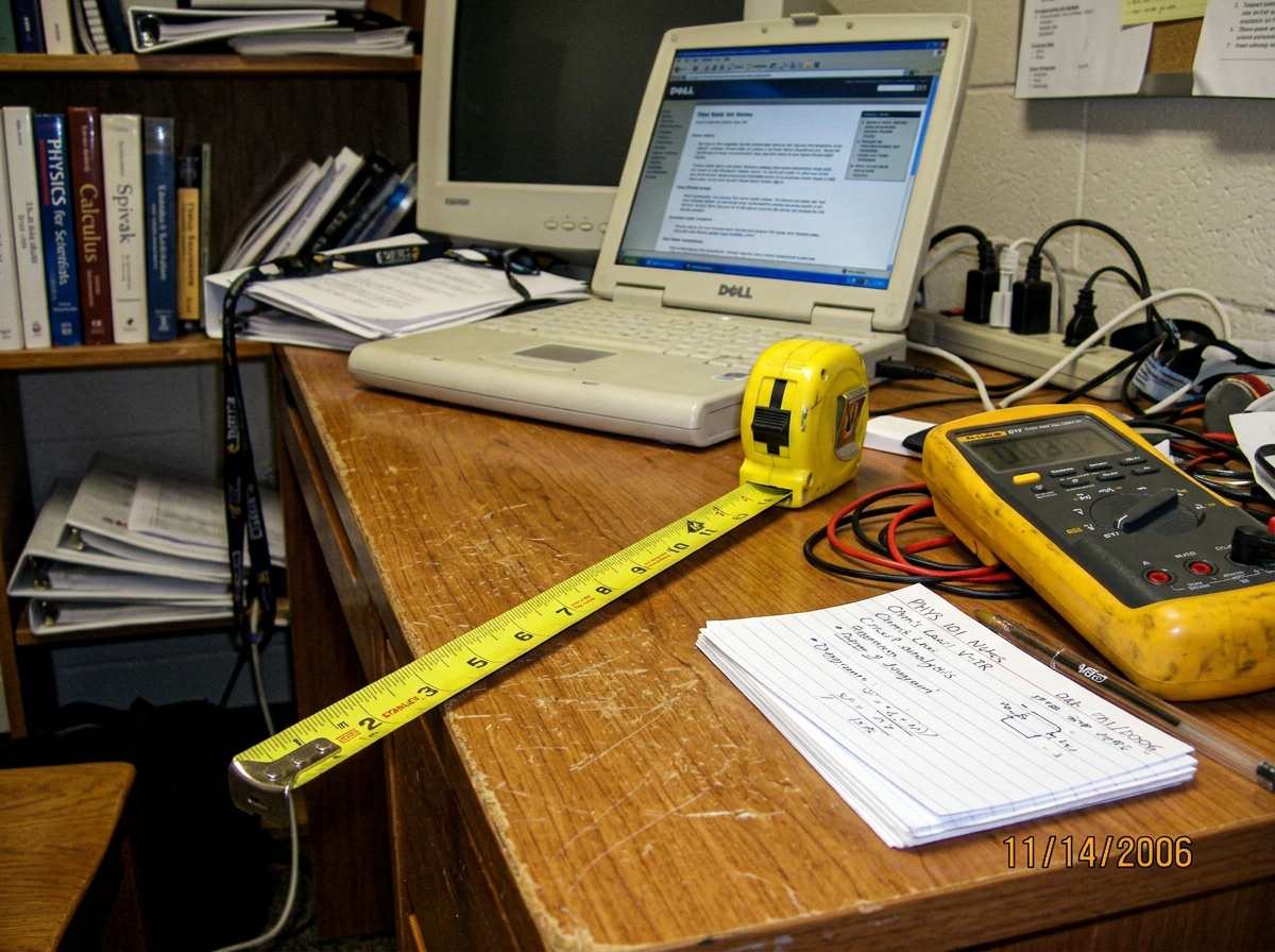 A yellow Stanley measuring tape partially unrolled across a dorm room desk, next to a Fluke 87V multimeter and a stack of index cards with neat handwriting. A close-up shot that emphasizes the contrast between the physical measuring tool and the academic clutter in the background.