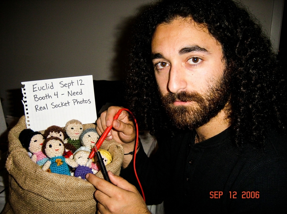 Digital multimeter probe resting next to a burlap sack of crocheted Gabe-ables dolls, with handwritten note reading 'Euclid Sept 12 Booth 4 - Need Real Socket Photos'