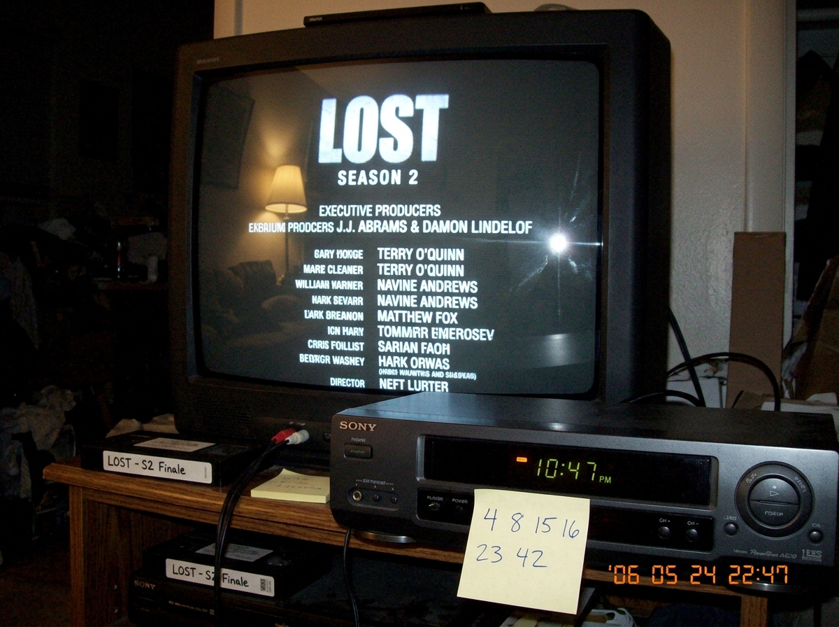 A blurry photo taken with a digital camera of a CRT television screen showing the end credits of Lost season 2. The screen has a slight glare from a lamp. On the VCR below the TV, the clock reads 10:47 PM. A handwritten sticky note on the VCR says '4 8 15 16 23 42' in blue pen.