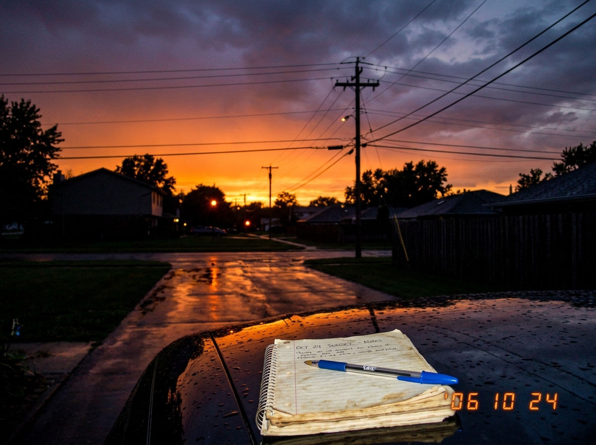 A photo taken from a suburban driveway looking west at the sky. Deep orange at the horizon fades into purple above. Power lines cross the frame. The driveway is wet from recent rain and reflects the orange color. In the foreground, the edge of a spiral notebook sits on a car hood with a blue pen res