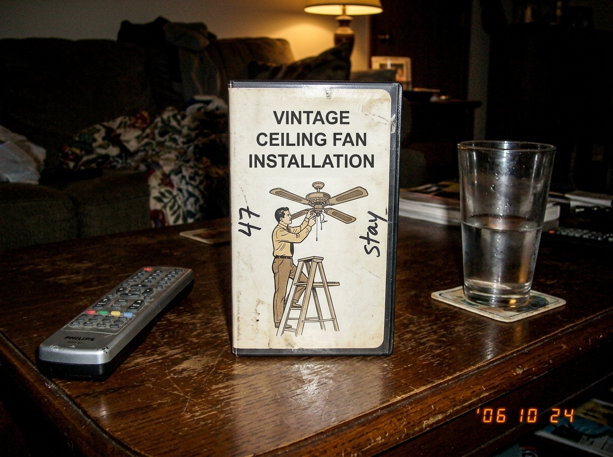 A photo of a vintage ceiling fan installation VHS tape on a coffee table. The cover has handwritten '47' and 'stay' scribbled in the margins in black marker. A remote control and a half-drunk glass of water sit next to it. The lighting is dim, yellowish from a single lamp.