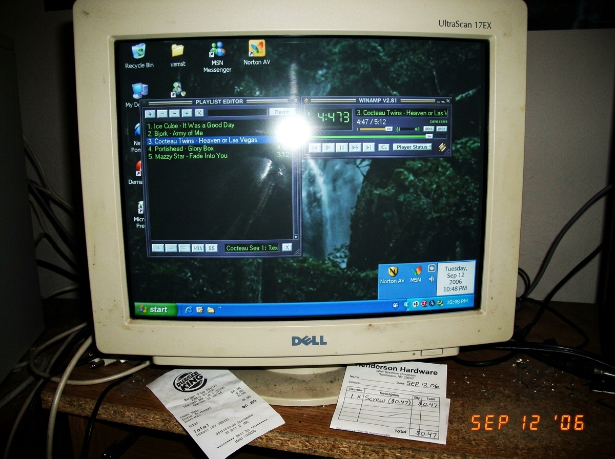 Blurry camera-phone photo of a CRT monitor showing a Winamp playlist. The track 'Heaven or Las Vegas' by Cocteau Twins is selected, and the Winamp timer displays 4:47. The system tray on the Windows desktop shows Sept 12 2006. On the desk beside the monitor, a crumpled fast-food receipt from a burge