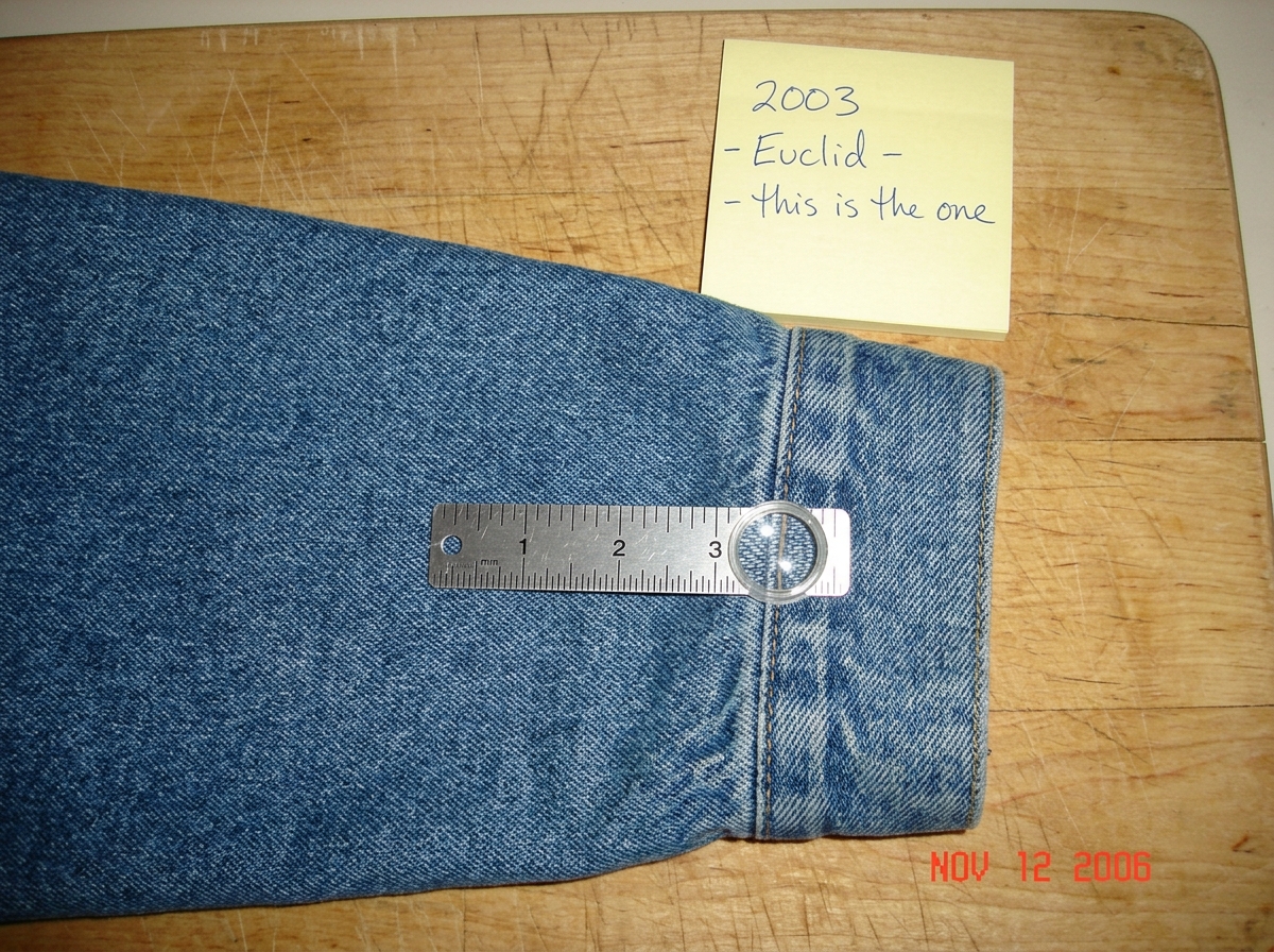 A close-up photo of a denim jacket cuff, held flat on a wooden cutting board. A silver sewing gauge with clearly marked inches and a magnifying lens sits on the fabric, aligned with the weave. The threads are visible at roughly 47 per inch if you count them carefully. A yellow Post-it note in the co