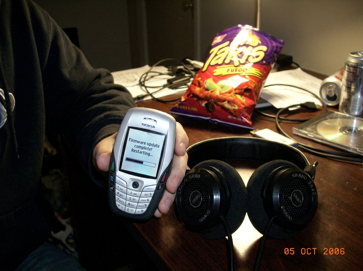 blurry camera phone photo of a Nokia 6600 held up next to a pair of Grado SR80 headphones on a messy bedroom desk. The Nokia screen shows a 'firmware update complete' message. A half-eaten bag of Takis is in the background.