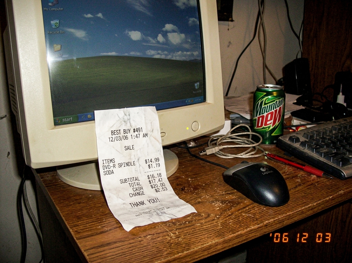 Low-quality digital camera photo of a crumpled paper receipt on a desk next to a CRT monitor, timestamp visible on receipt reads 1:47 AM
