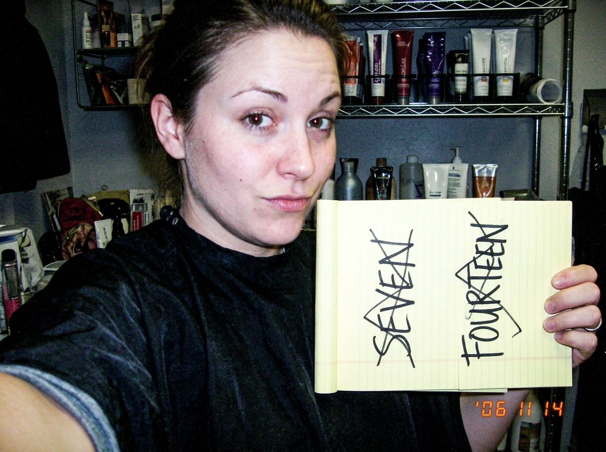 A grainy webcam selfie of a woman with a salon smock, clearly taken in a staff-only back room. She holds a yellow legal pad with two columns written in black marker. Left column: SEVEN. Right column: FOURTEEN. Both are crossed out with angry zigzag lines. In the background, partially visible on a sh