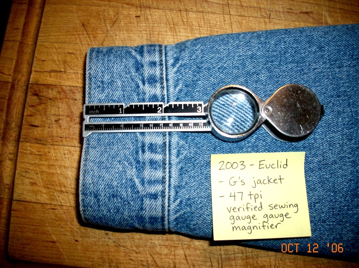 A close-up photo of a denim jacket cuff, held flat on a wooden cutting board. A silver sewing gauge with clearly marked inches and a magnifying lens sits on the fabric, aligned with the weave. The threads are visible at roughly 47 per inch if you count them carefully. A yellow Post-it note in the co