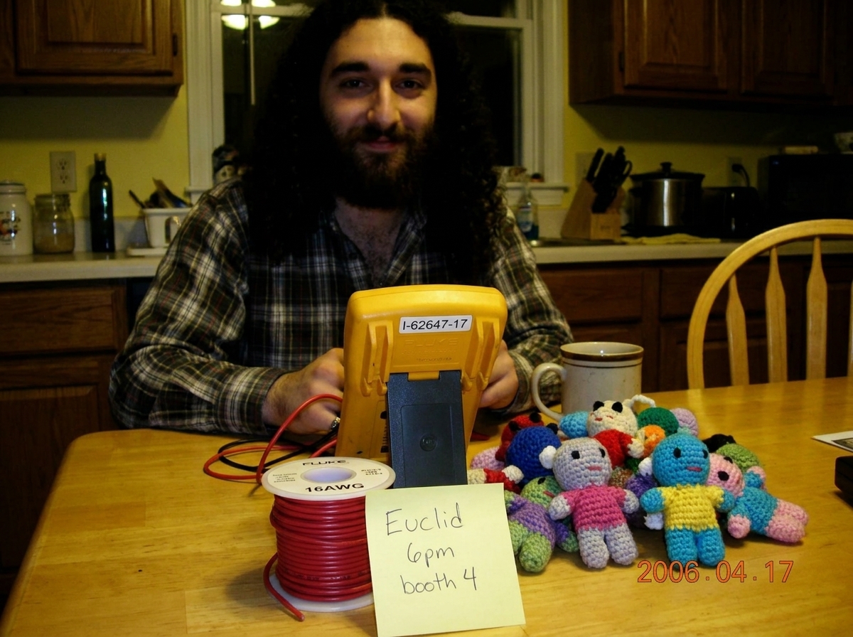 Photo of a Fluke 87V multimeter on a kitchen table, serial number I-62647-17 visible on the back, next to a spool of 16awg wire and a small pile of crocheted Gabe-ables plush toys. A yellow Post-it note reads 'Euclid 6pm booth 4' and a coffee mug sits nearby.