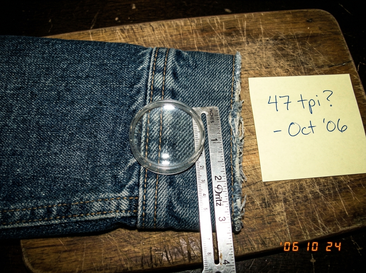 A close-up photo of a denim jacket cuff, held flat on a wooden cutting board with a silver sewing gauge showing inches and a magnifying lens, aligned with the weave. The threads are visible at roughly 47 per inch. A yellow Post-it note is nearby.