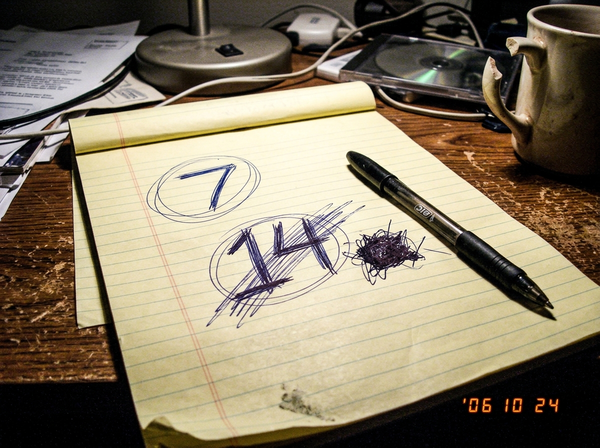 A legal pad photo taken at a messy desk under a lamp. On the paper, the numbers 7 and 14 are scrawled and circled, with aggressive cross-outs through the 14. A pen sits on the pad and a coffee mug with a broken handle is visible in the corner.