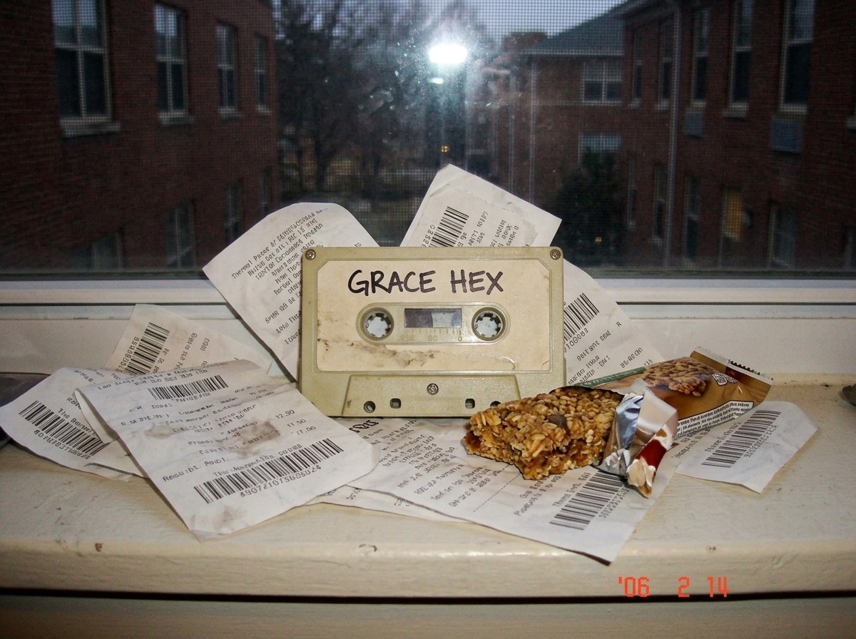 Blurry phone photo of a beige GRACE HEX cassette tape sitting on a dorm windowsill, surrounded by crumpled thermal paper receipts with gamma scan codes arranged like a shrine, next to a half-eaten stale granola bar
