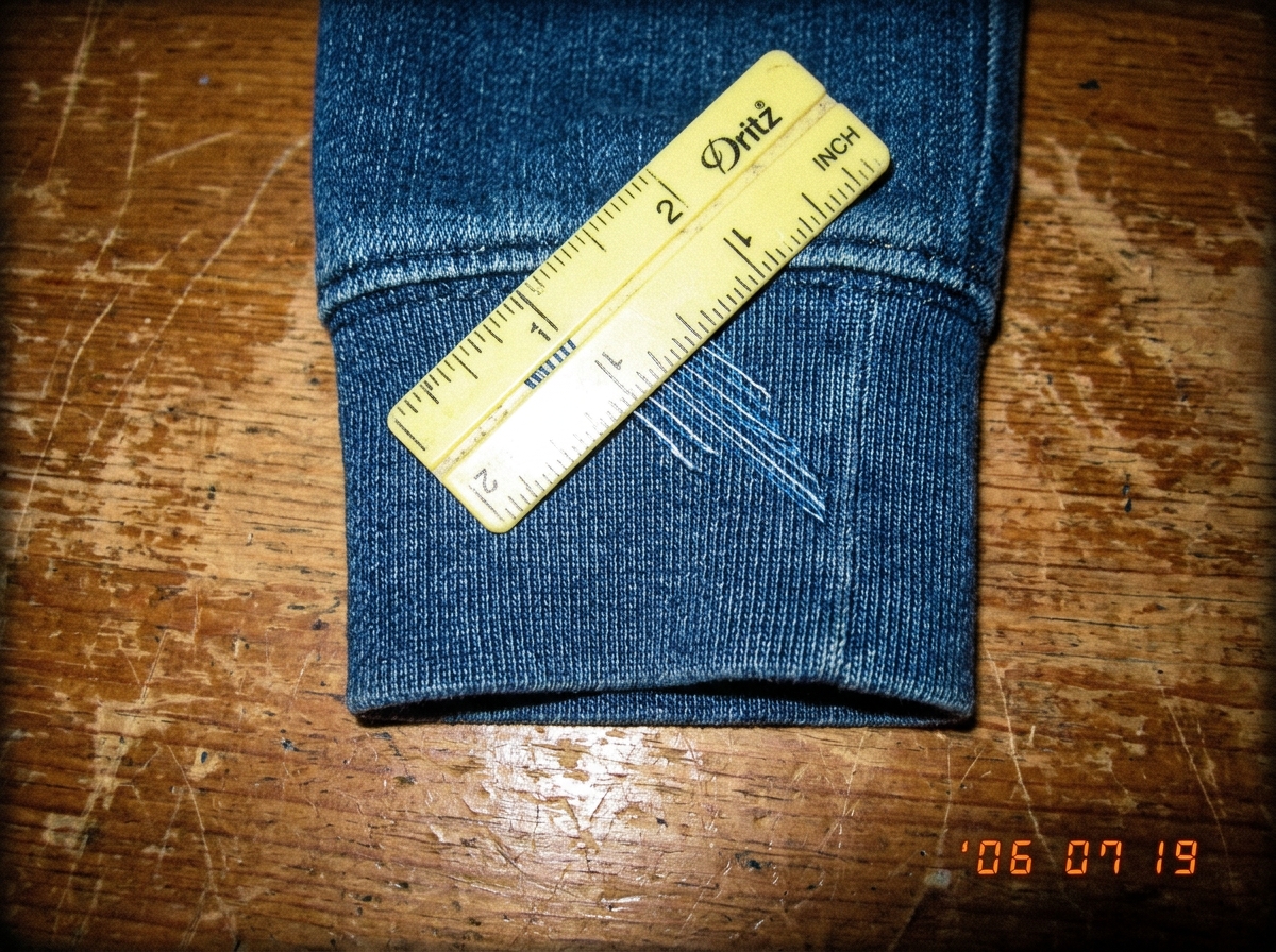 A close-up photograph of a denim hoodie cuff on a wooden table, with a yellow plastic sewing gauge laid across the fabric measuring the thread count. The gauge shows approximately 47 threads per inch. Natural window light. The photo is slightly blurry at the edges, taken with an early 2000s digital 