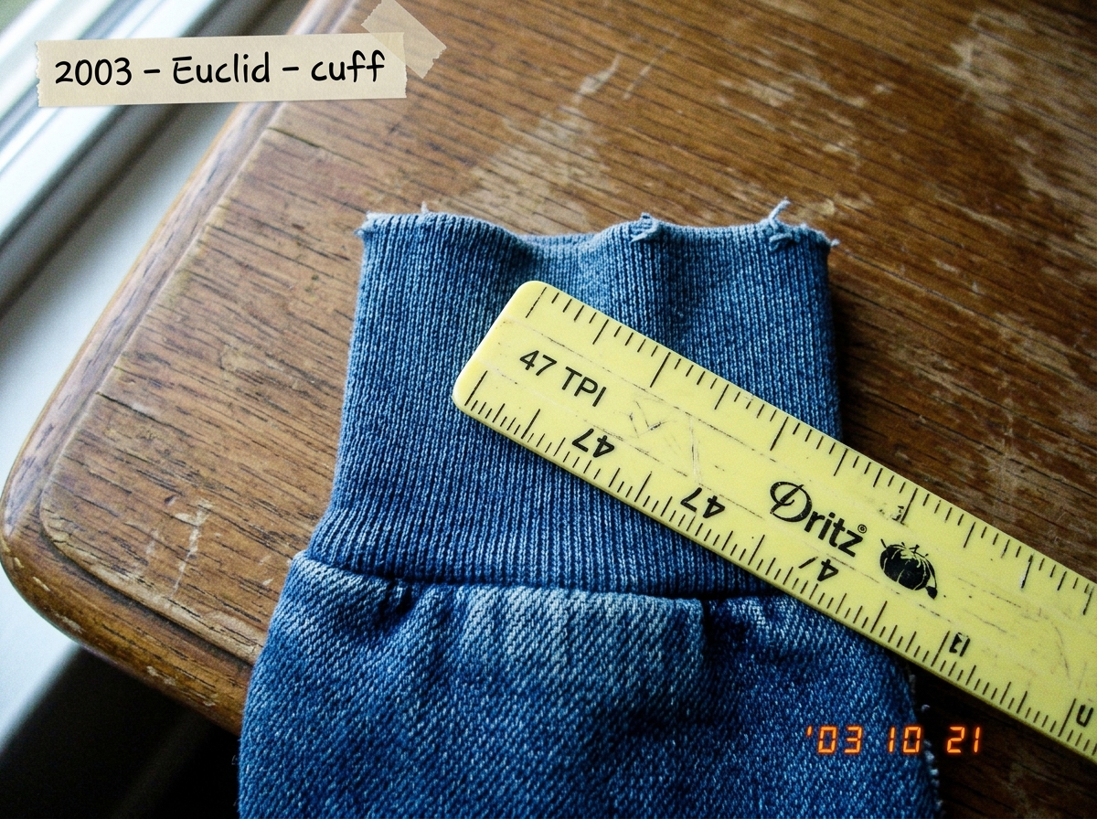 A close-up photograph of a denim hoodie cuff on a wooden table, with a yellow plastic sewing gauge laid across the fabric measuring the thread count. The gauge shows approximately 47 threads per inch. Natural window light. The photo is slightly blurry at the edges, taken with an early 2000s digital 