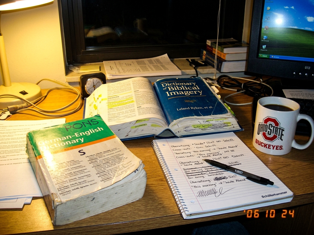 A photo of a desk at night: a German-English dictionary open to the letter 'S', a blue hardcover 'Dictionary of Biblical Imagery' open beside it, a notepad with handwritten cross-outs in German and English, and a coffee mug with Ohio State logo. Low lamp light, messy but organized.