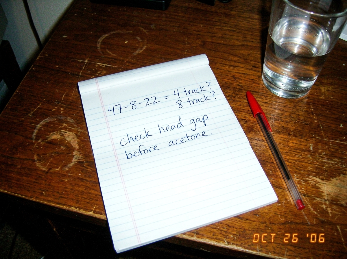 A close-up photo taken with a low-res 2006 digital camera showing a piece of notebook paper. On it, handwritten in blue ballpoint pen: '47-8-22 = 4 track? 8 track? check head gap before acetone.' The handwriting is messy, slightly tilted. The paper is on a dark wooden desk next to a half-empty glass