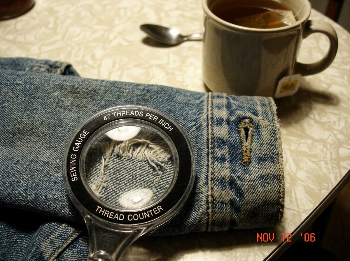 A close-up photo of a worn denim jacket cuff, with a sewing gauge magnifier laid over the fabric showing threads counted at 47 per inch, on a kitchen table with a cup of tea nearby