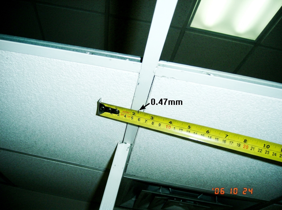 A close-up photograph of a ceiling tile grid showing a measured gap of 0.47mm between two white acoustic tiles. A yellow tape measure lies across the seam, with the 0.47mm mark visible. The ceiling is in a commercial room with fluorescent lighting visible in the corner.