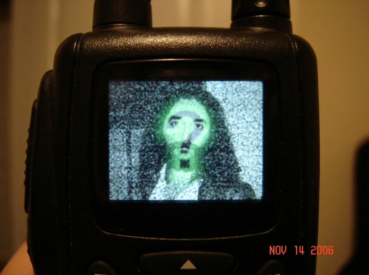 A close-up of a vintage walkie-talkie's static screen with a faint spectral green glow forming the shape of a question mark, subtly resembling Gabriel Chantayan's silhouette