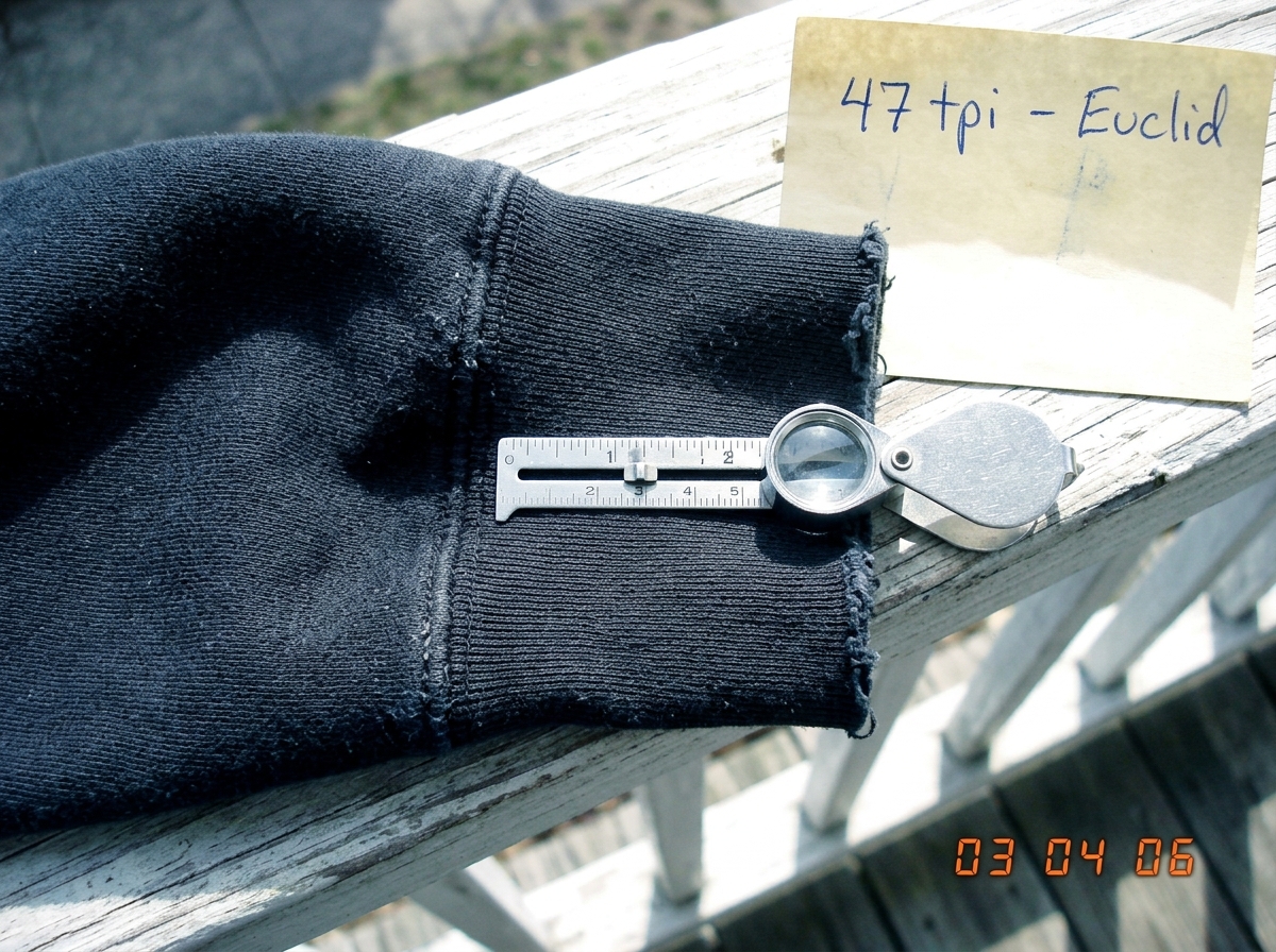 A close-up photograph of a dark hoodie cuff taken in 2003, laid flat on a wooden porch railing in daylight. The cuff fabric is a heavy cotton sweatshirt material, visibly worn at the edge. A silver sewing gauge with a magnifying lens is placed across the weave, showing thread count measurement. A ha