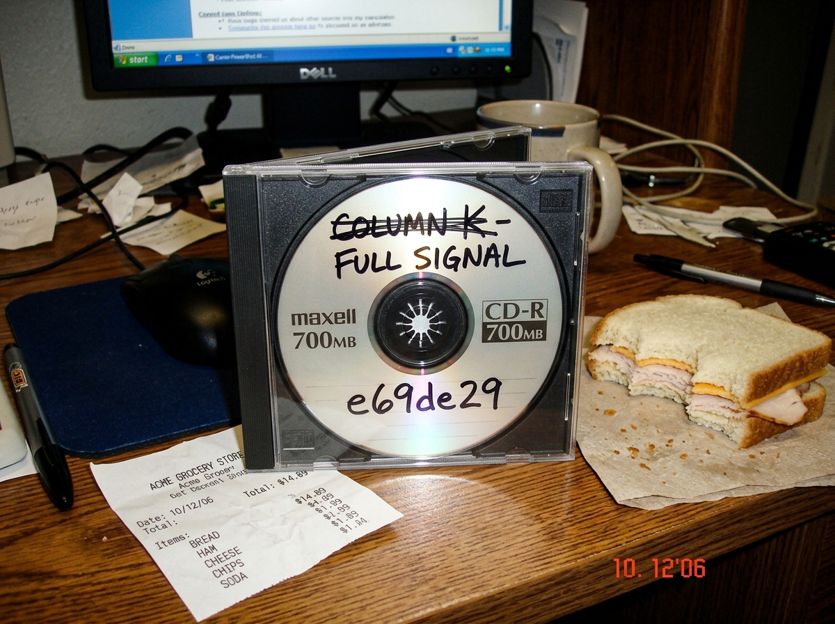 A photograph taken at a slight angle on a messy desk. A Maxell CD-R disc in a slim jewel case sits next to a receipt from Acme grocery. On the CD-R surface, someone has written in black marker 'COLUMN K - FULL SIGNAL' and then crossed it out and written 'e69de29' underneath. Beside the jewel case is