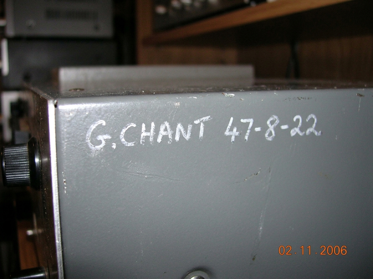 A close-up of a reel-to-reel tape recorder's metal box showing 'G.CHANT 47-8-22' written in faint white grease pencil, slightly smudged. The lighting is warm and slightly dim, emphasizing the texture of the metal and the pencil marks. The background is blurred, focusing only on the writing.