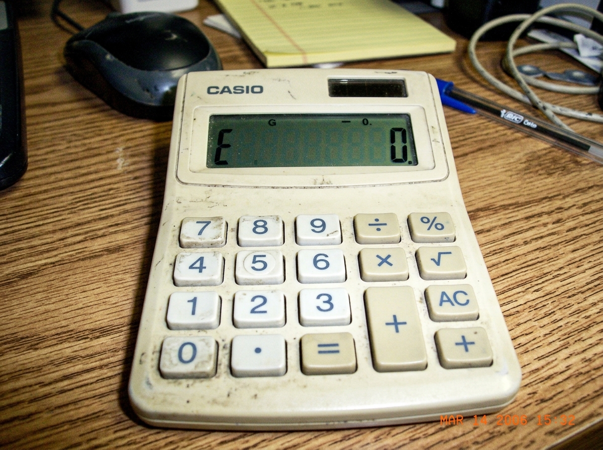 A screenshot of a basic four-function calculator showing 'ERROR' after attempting to divide by zero. The display reads 'E' on a small LCD screen. The calculator is a standard beige desktop model with visible buttons.