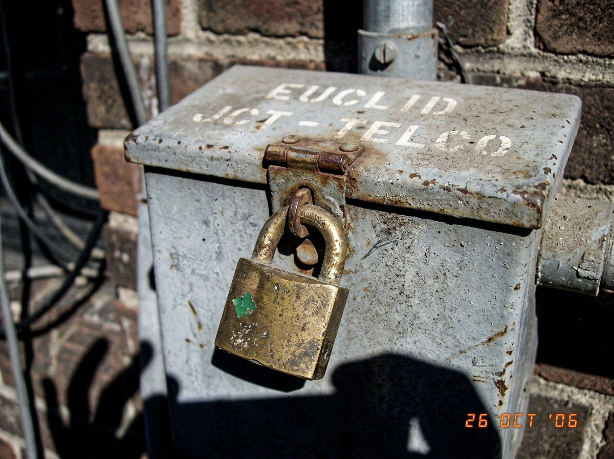A close-up of a brass padlock on a metal junction box. The padlock is dusty and tarnished, with a green paint fleck visible on one side. The junction box is labelled 'EUCLID JCT - TELCO' in faded stencilled letters. Hand shadow visible at the edge of frame from the photographer holding the camera.