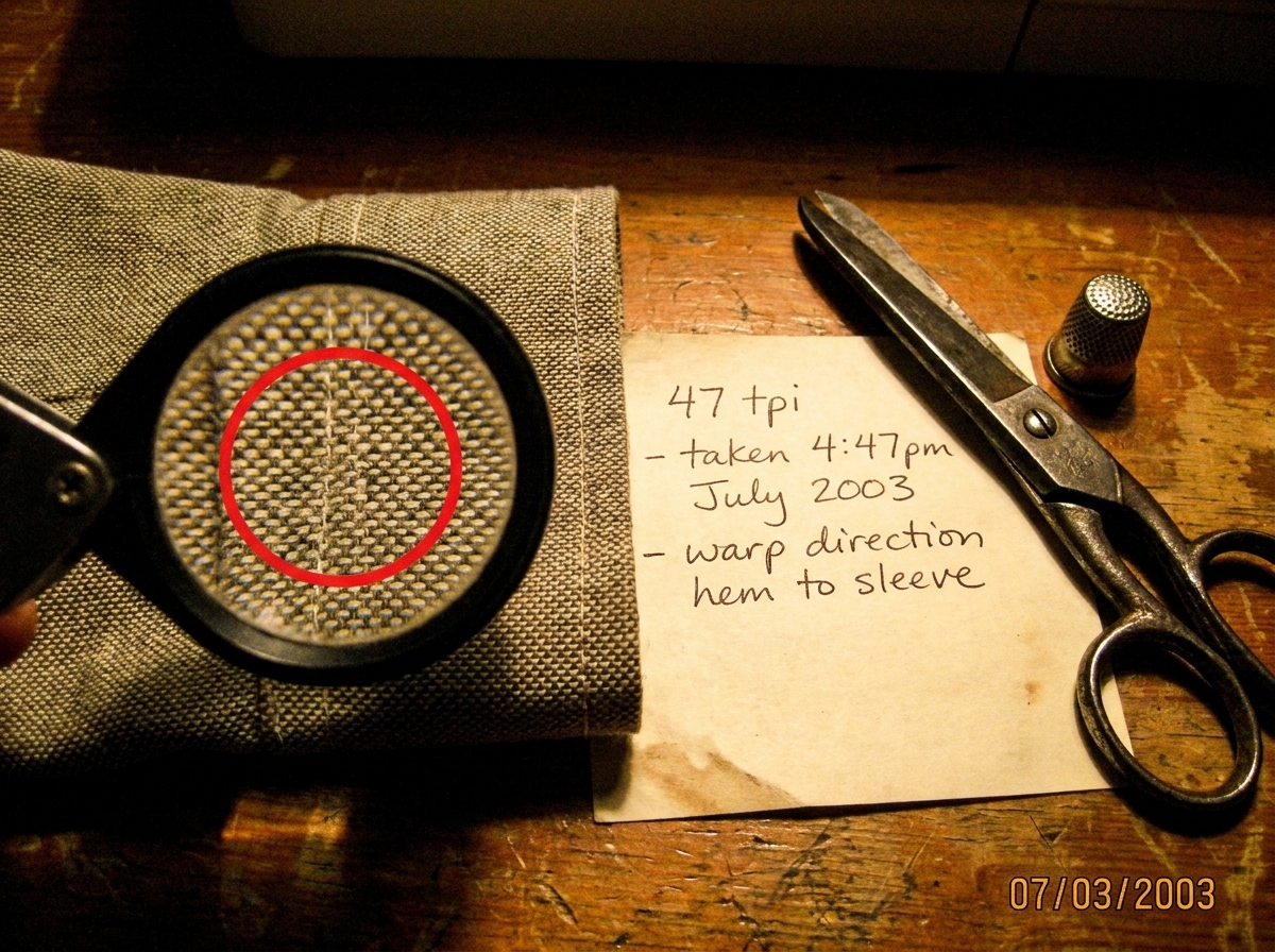 A close-up macro photo of a woven fabric cuff, taken with a sewing gauge magnifier showing the weave. A red circle highlights the thread count area. A handwritten note in the corner reads '47 tpi - taken 4:47pm July 2003 - warp direction hem to sleeve'. The photo is slightly yellowed, taken on a woo