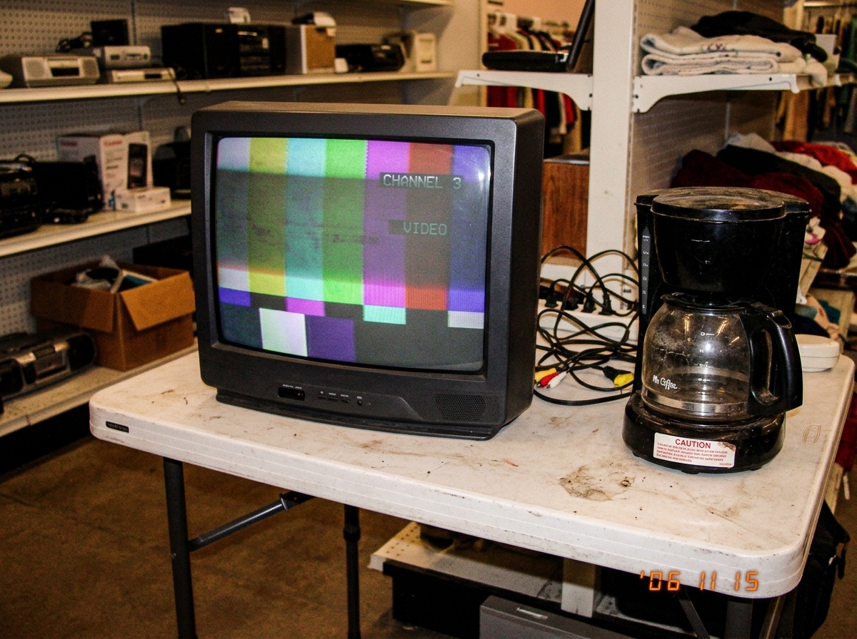 blurry phone photo of a thrift store CRT monitor displaying a dim green and purple test pattern with visible screen burn, sitting on a wobbly plastic table next to a coffee maker with a 'CAUTION' sticker