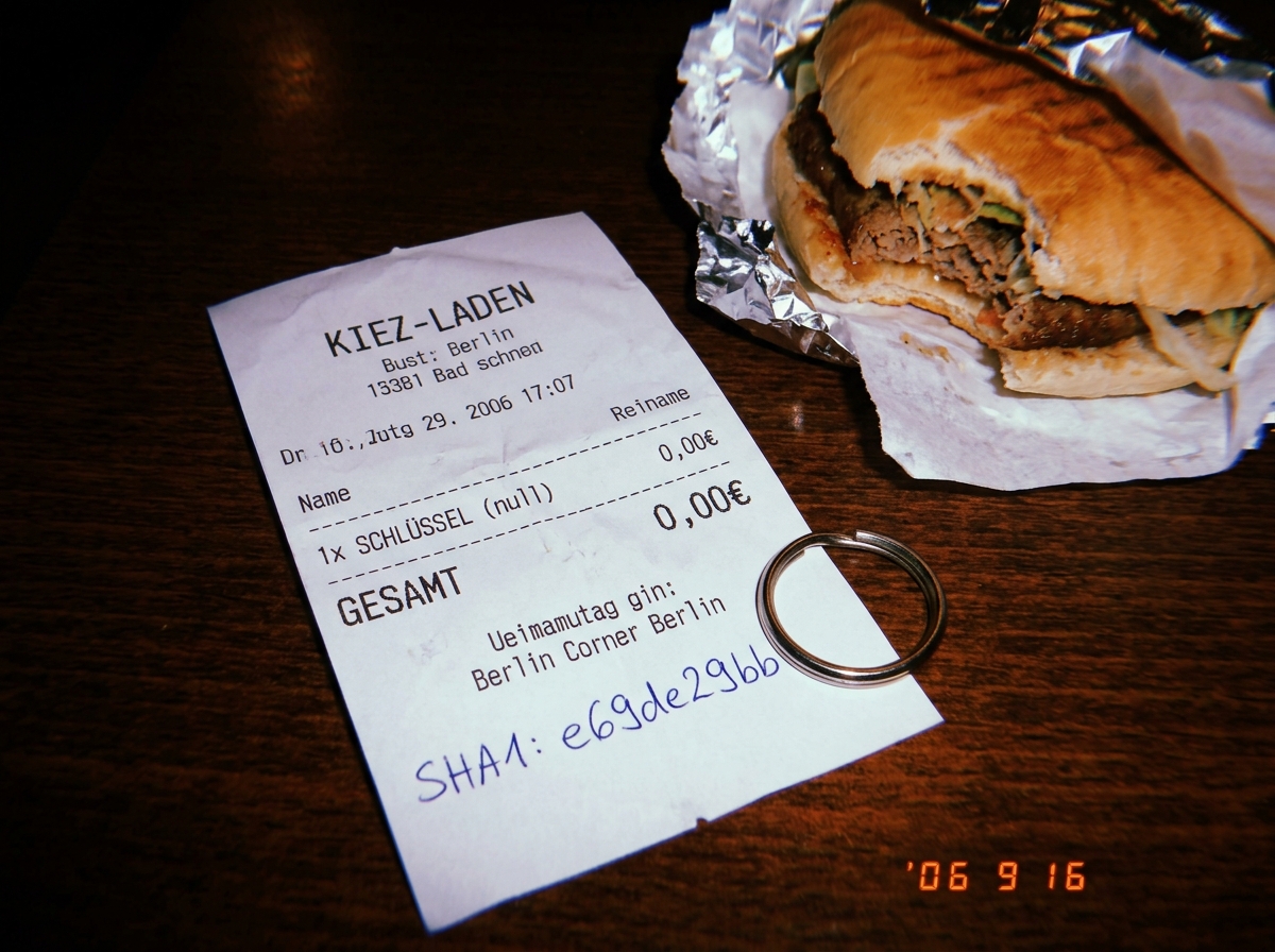 A crumpled receipt from a Berlin corner shop, dated 16.09.06, listing only '1x SCHLÜSSEL (null)' with a total of 0,00€. The bottom has a handwritten SHA1 hash: e69de29bb. The receipt is photographed on a dark wooden table next to a half-eaten döner and a keyring with no keys.