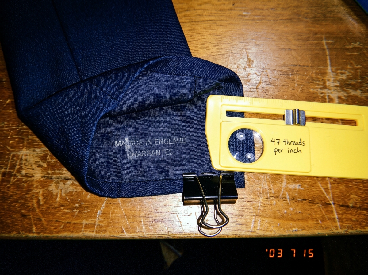 A close-up photograph of a dark blue woven fabric cuff, perhaps from a jacket sleeve, laid flat on a wooden table next to a yellow plastic sewing gauge. The gauge has a magnifier and the measurement markings are visible, showing 47 threads per inch. The cuff has a faint watermark stamp on the inner 