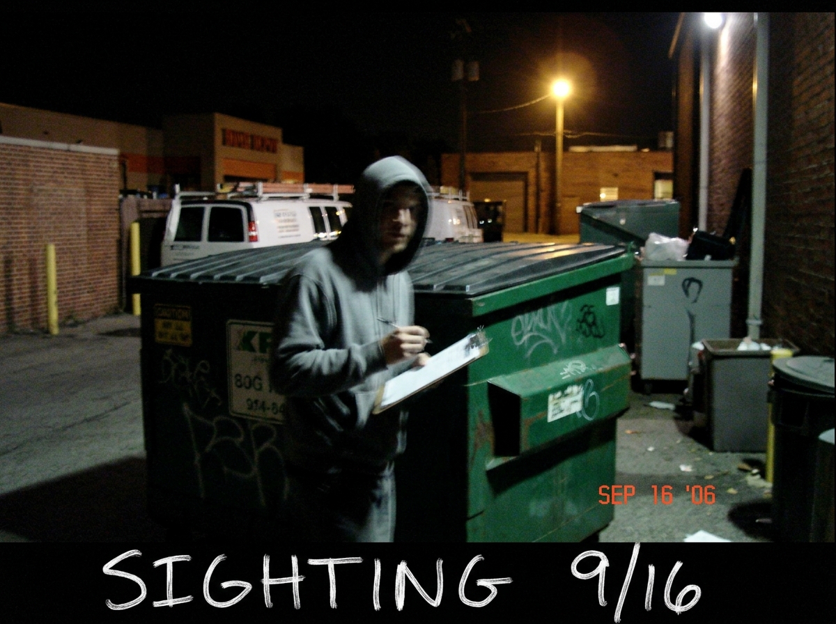 A grainy, low-resolution digital photo taken at night showing a shadowy figure in a grey hoodie standing next to a dumpster behind a Home Depot. The figure is holding a clipboard. The text 'SIGHTING 9/16' is handwritten on the bottom of the photo in white marker.