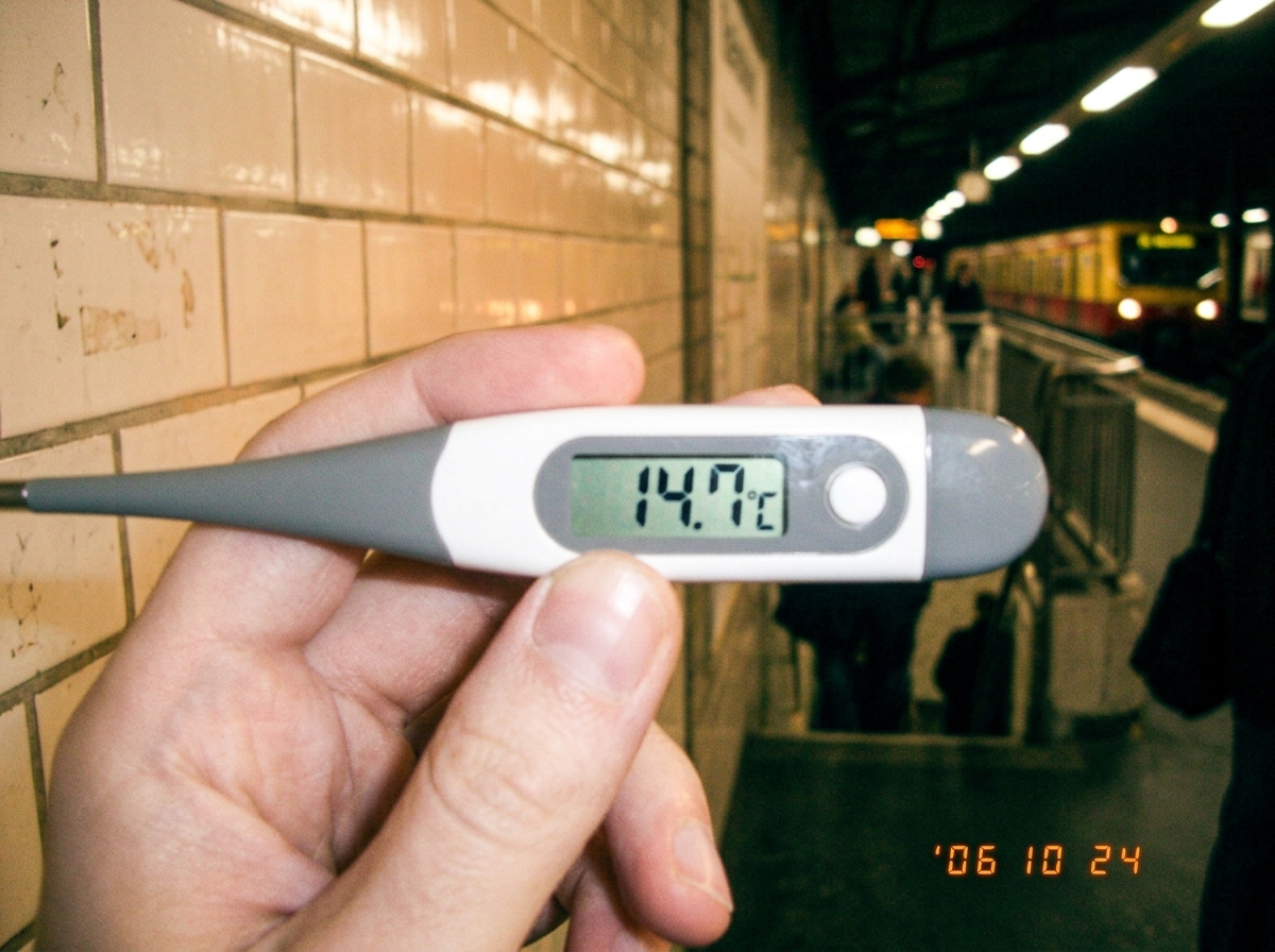 A digital thermometer reading 14.7°C held in front of a train station platform tile showing a faint warm glow on the surface, evening light