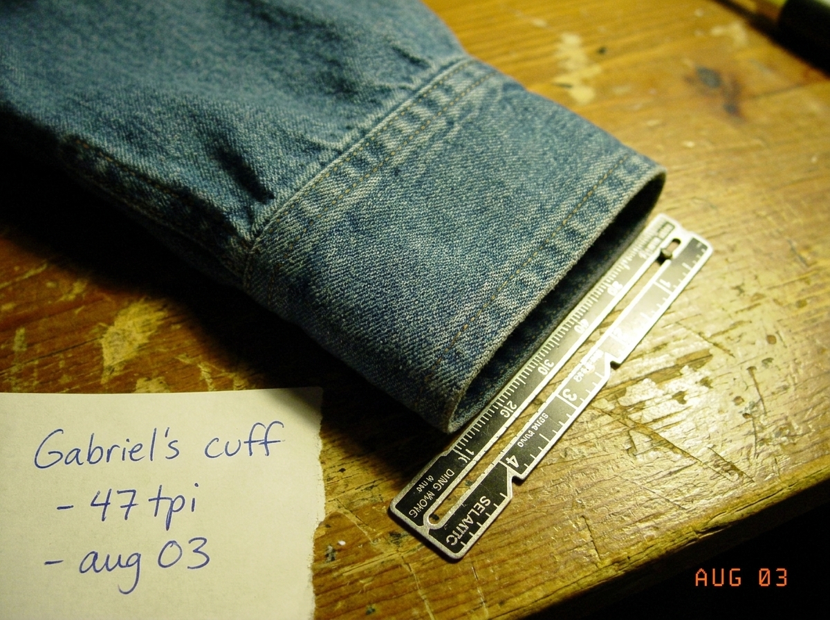 A close-up photo of a denim shirt cuff resting on a wooden table, visible weave pattern with a sewing gauge ruler placed next to it measuring approximately 47 threads per inch. The photo is slightly out of focus and has a yellowed tint suggesting it was taken in 2003. A handwritten note in the corne