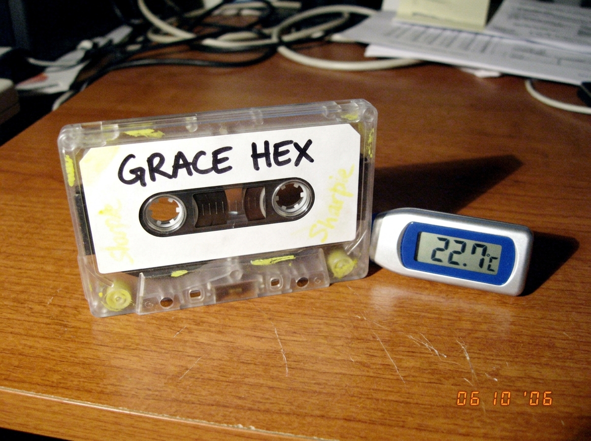 Cassette tape labeled GRACE HEX sitting on a desk next to a digital thermometer showing 22.7°C, with faint yellow sharpie smudges visible on the tape casing