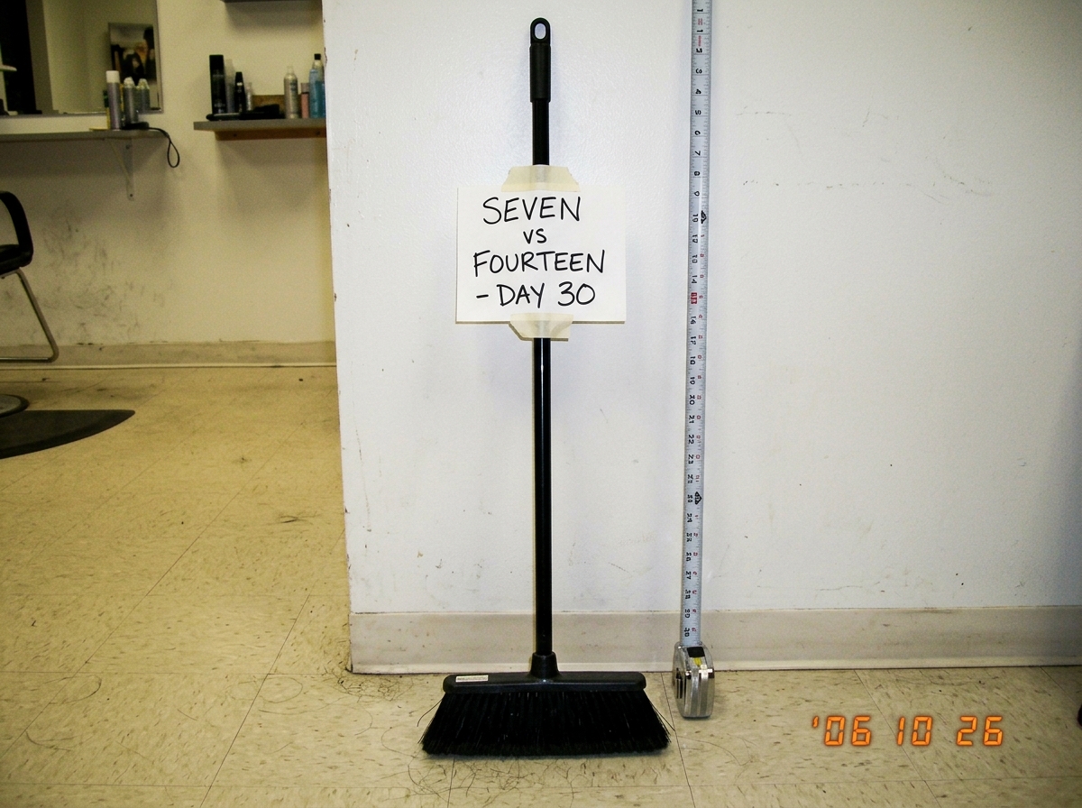 A professional salon broom standing upright against a white wall, with a handwritten sign taped to its handle reading 'SEVEN vs FOURTEEN - DAY 30' in black marker. The floor beneath it is clean and swept. A measuring tape is visible next to it for scale.