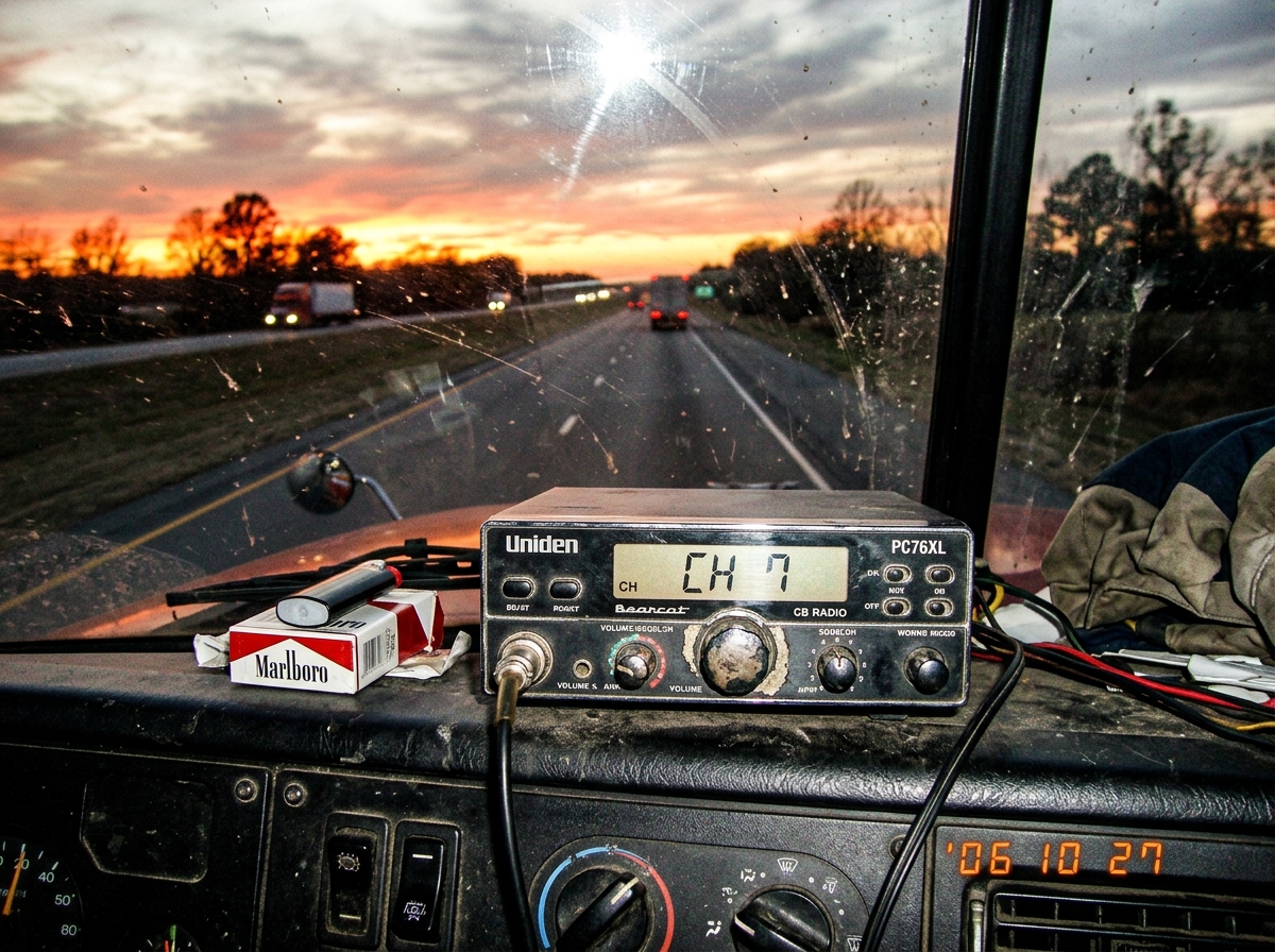 A close-up photo taken through a dusty truck windshield showing a CB radio mounted on the dashboard. The display shows channel 7, squelch turned to a specific point. Beyond the windshield, the sun is setting over a long straight highway, casting orange light across the cab. A pack of smokes sits nex